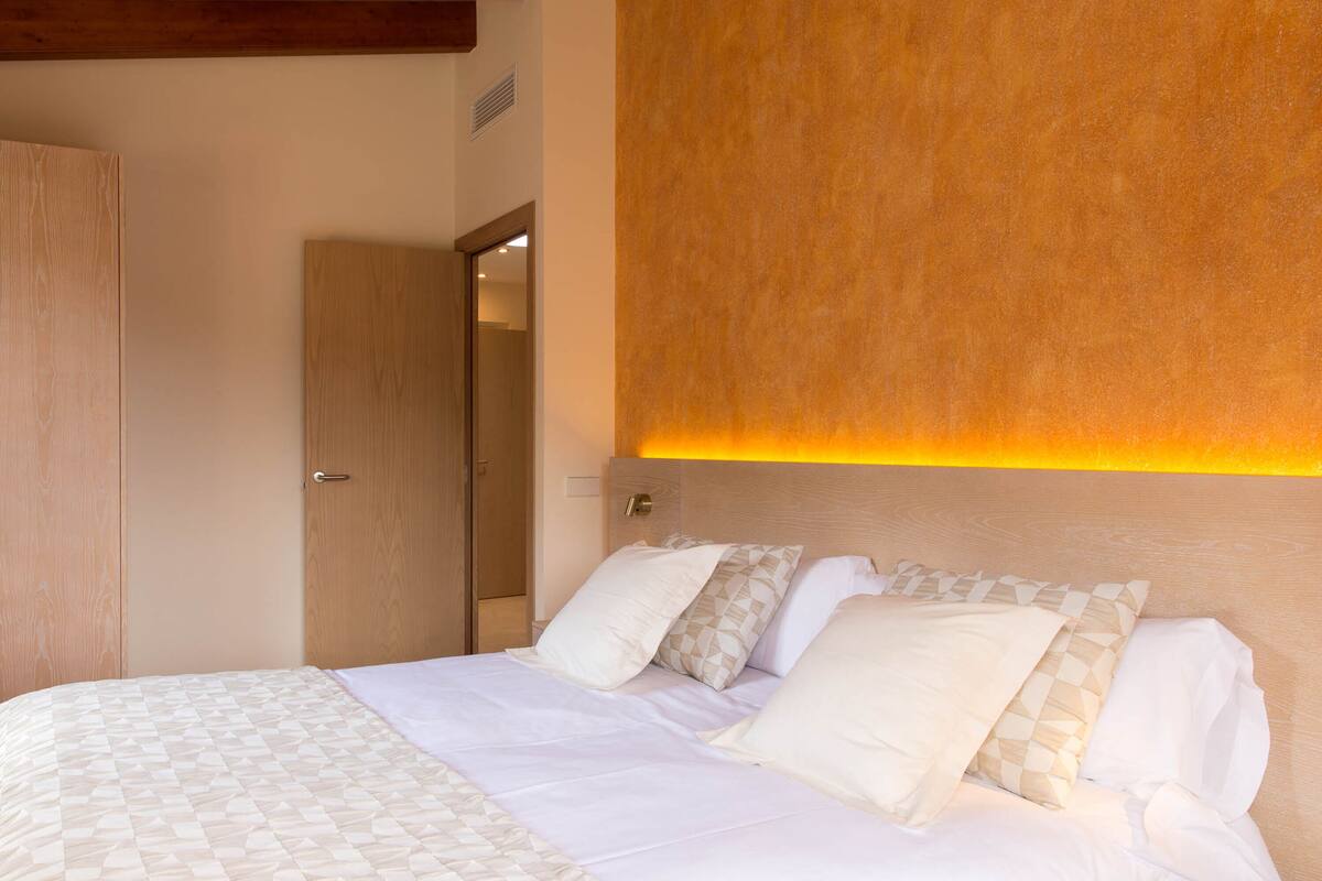 A neatly made bed with a patterned bedspread, a wooden headboard, and a wall-mounted light fixture.