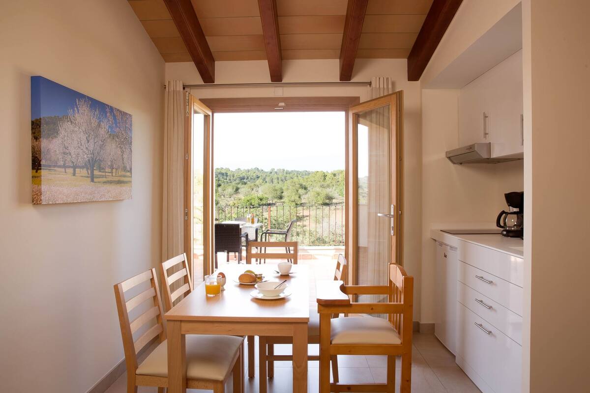 A dining area with a table set for two, a kitchen with white cabinets, and a view of a green landscape through an open door.