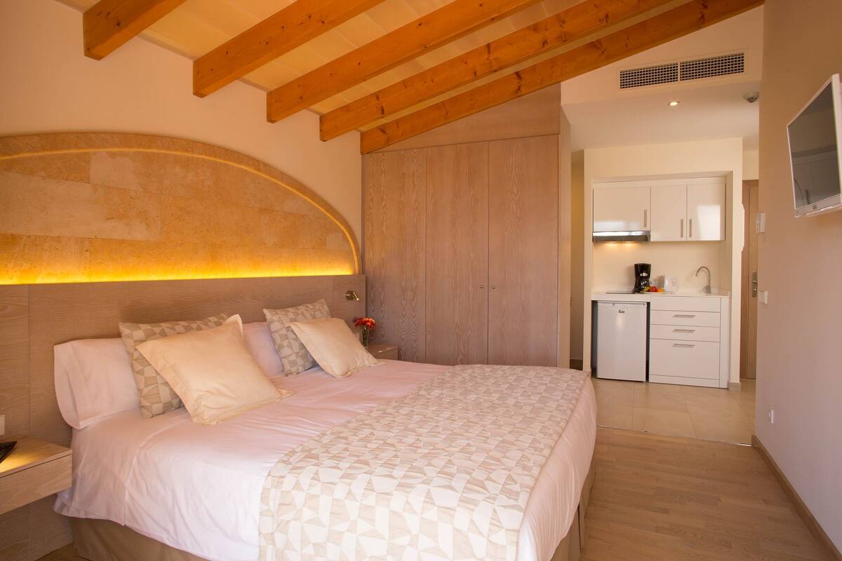 A bedroom with a large bed, wooden ceiling beams, a built-in wardrobe, and a kitchenette in the adjacent room.