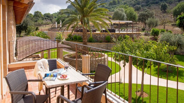 A balcony with wicker furniture, a table with a magazine and a glass, and a view of a landscaped garden with a palm tree.