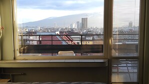 Una habitación con un ventanal que da a vistas de la ciudad, una cama con colcha y una silla junto a la ventana.
