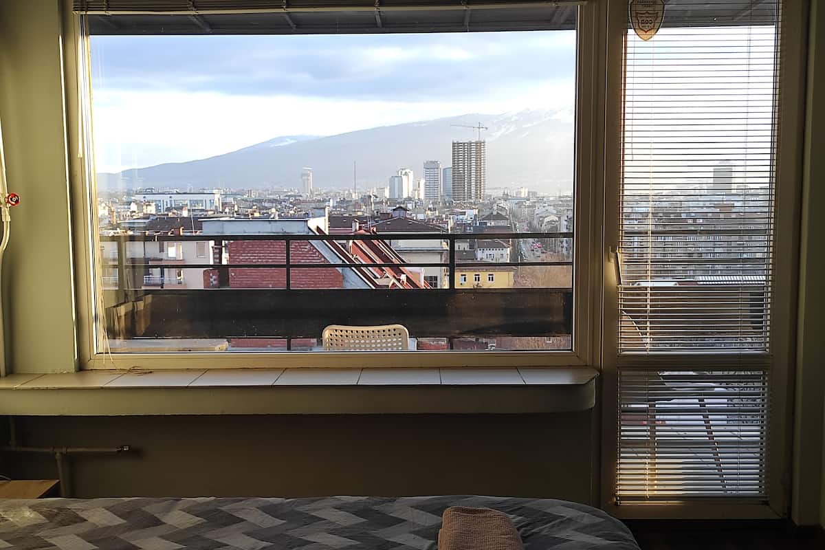 A room with a large window overlooking a cityscape, a bed with a quilt, and a chair by the window.
