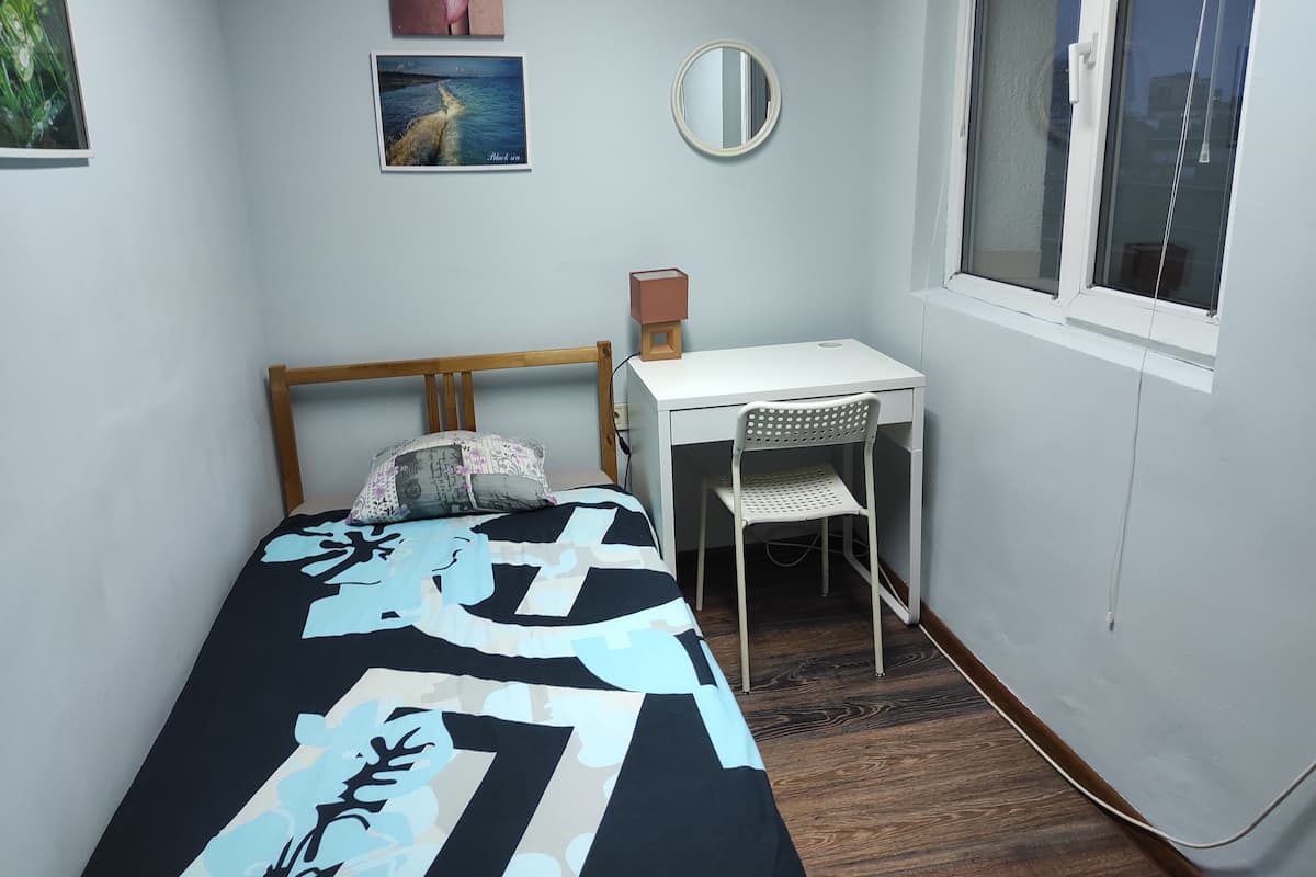 A small bedroom with a bed, a desk, a chair, a mirror, and a window.