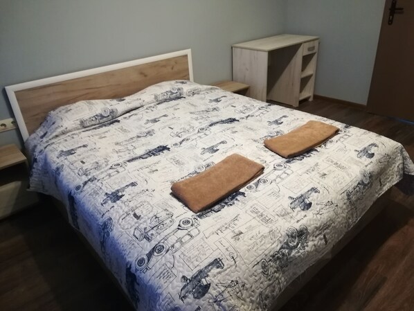 Una cama con una colcha estampada, dos toallas dobladas, un cabecero de madera, una mesita de noche y un pequeño armario de madera.