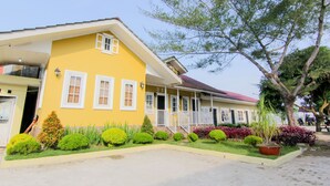 A yellow house with a red roof, white windows, and a porch. There are green bushes and a tree in front. - Trans Bandara Residence by Zuzu (Deli Serdang)