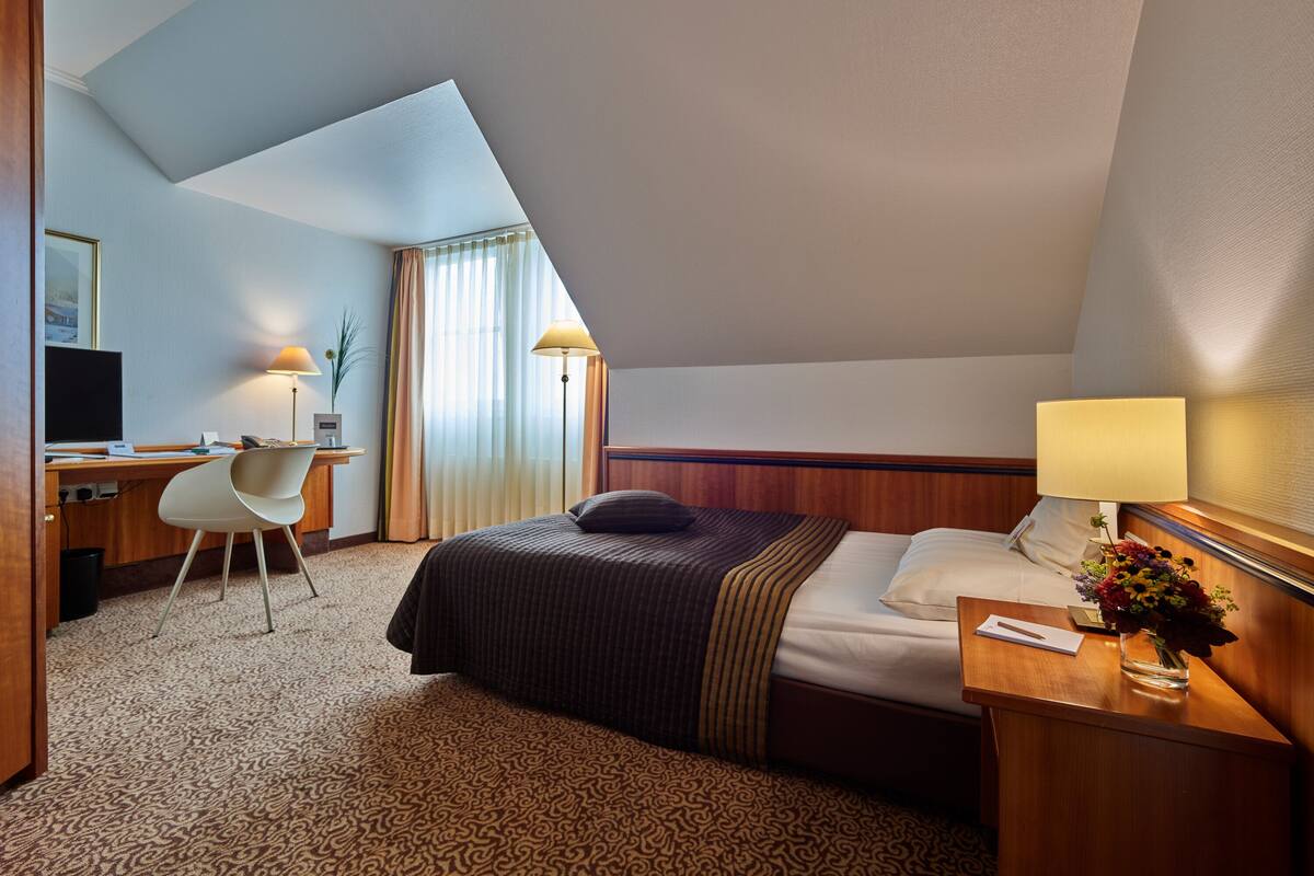 A hotel room with a bed, desk, chair, and a desk lamp.