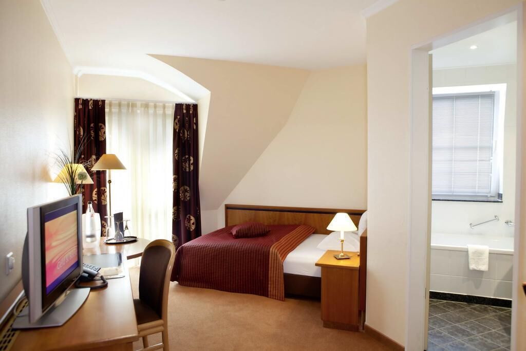 Habitación de hotel con cama, mesita de noche, lámpara, escritorio con computadora, silla y vista al baño.