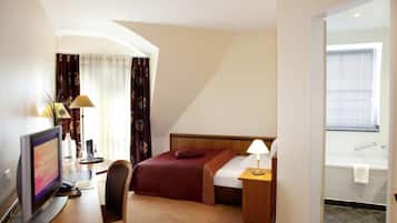 Habitación de hotel con cama, mesita de noche, lámpara, escritorio con computadora, silla y vista al baño.