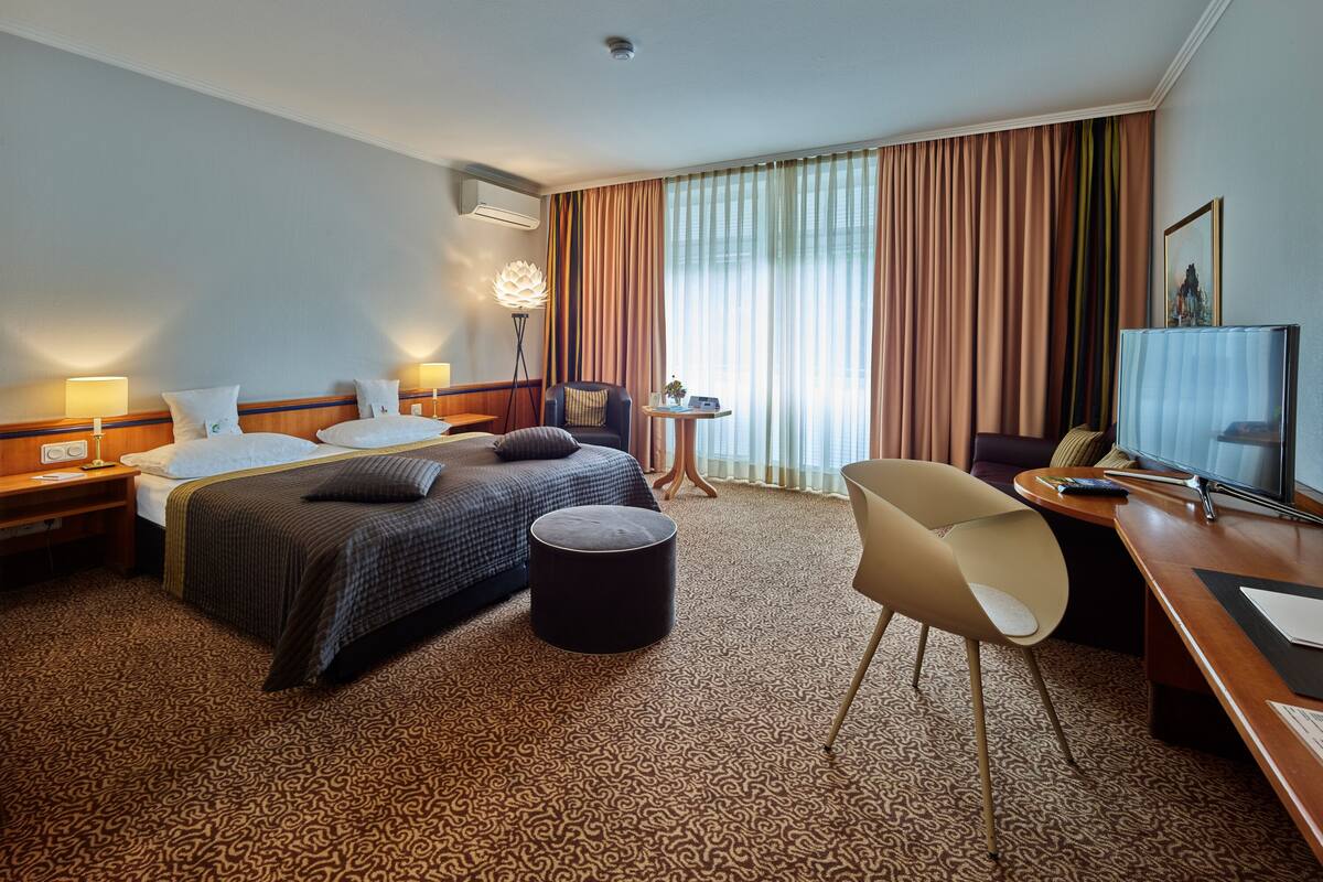 A hotel room with a bed, bedside tables, a desk with a chair, a lamp, a TV, and a window with curtains.