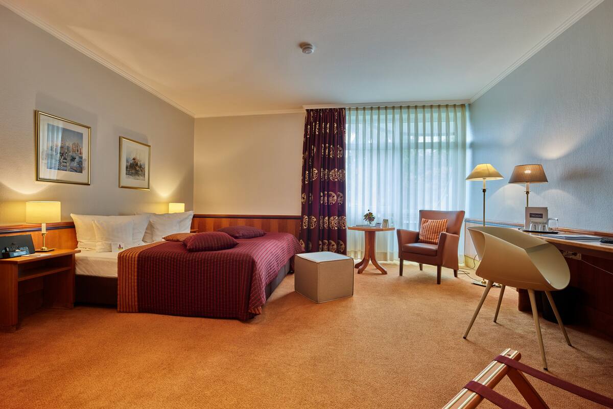 A hotel room with a bed, a desk, a chair, a small table, and a window with curtains.