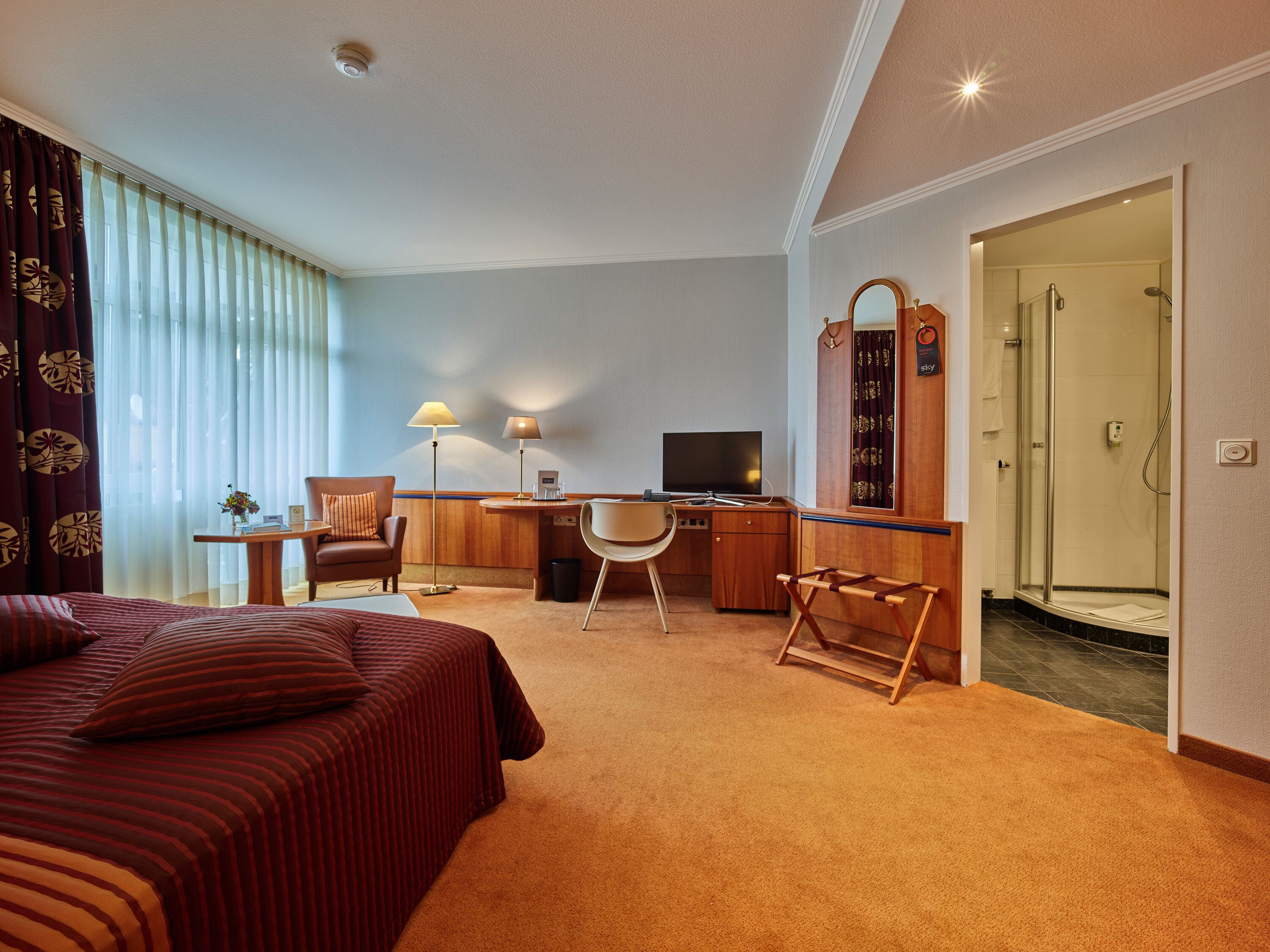 Ett hotellrum med en säng, ett skrivbord, en stol, en TV och en duschhörna.
