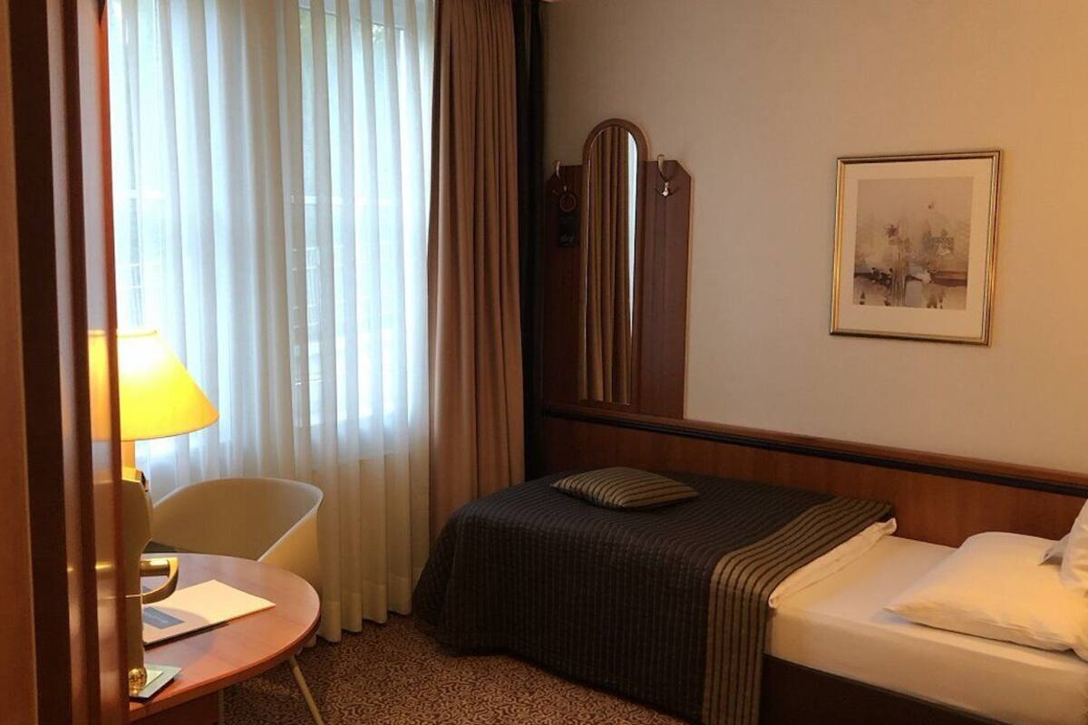 A hotel room with a bed, a desk with a lamp, a chair, and a framed picture on the wall.