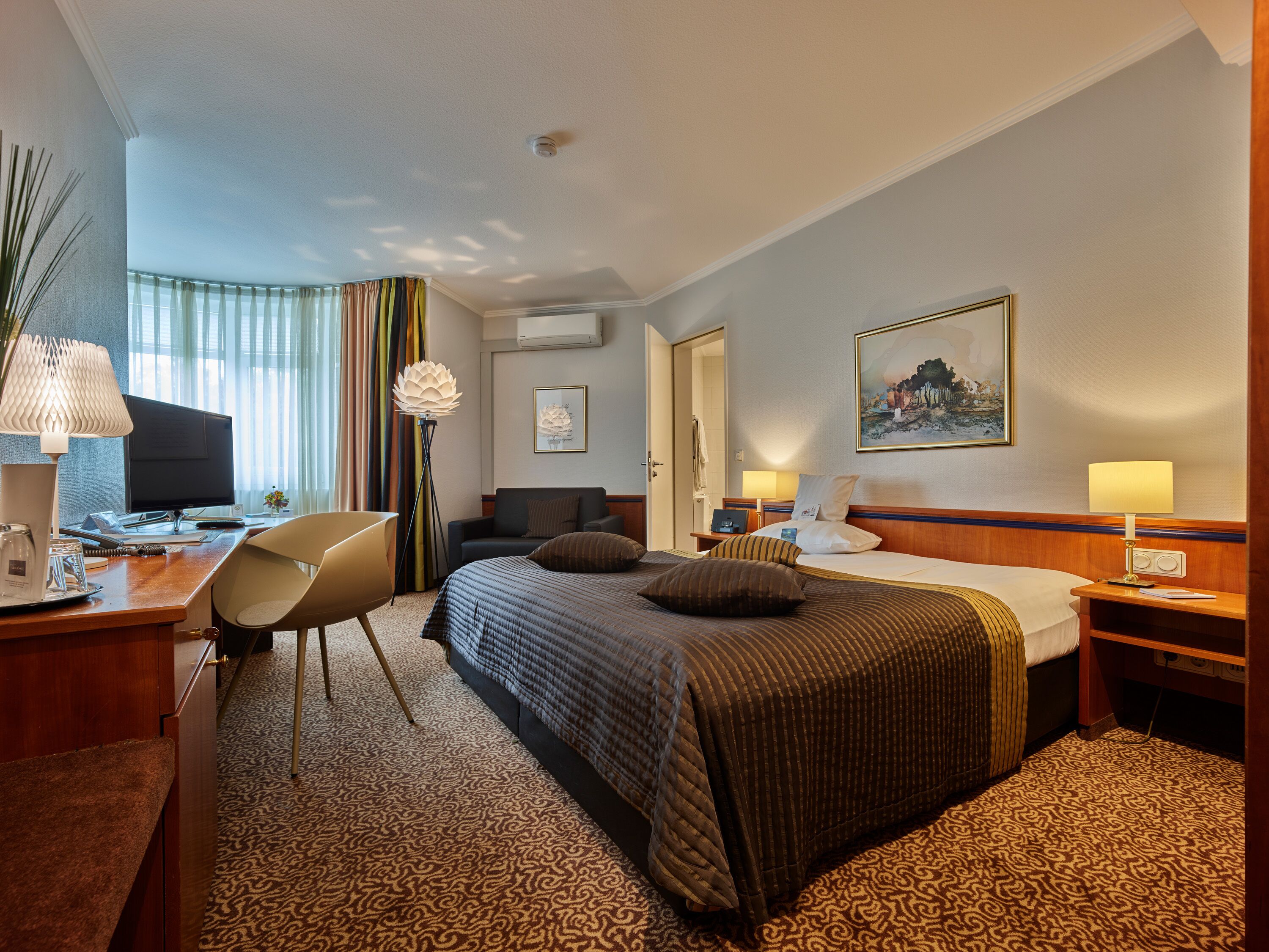 A hotel room with a bed, desk, chair, and a large window with curtains.