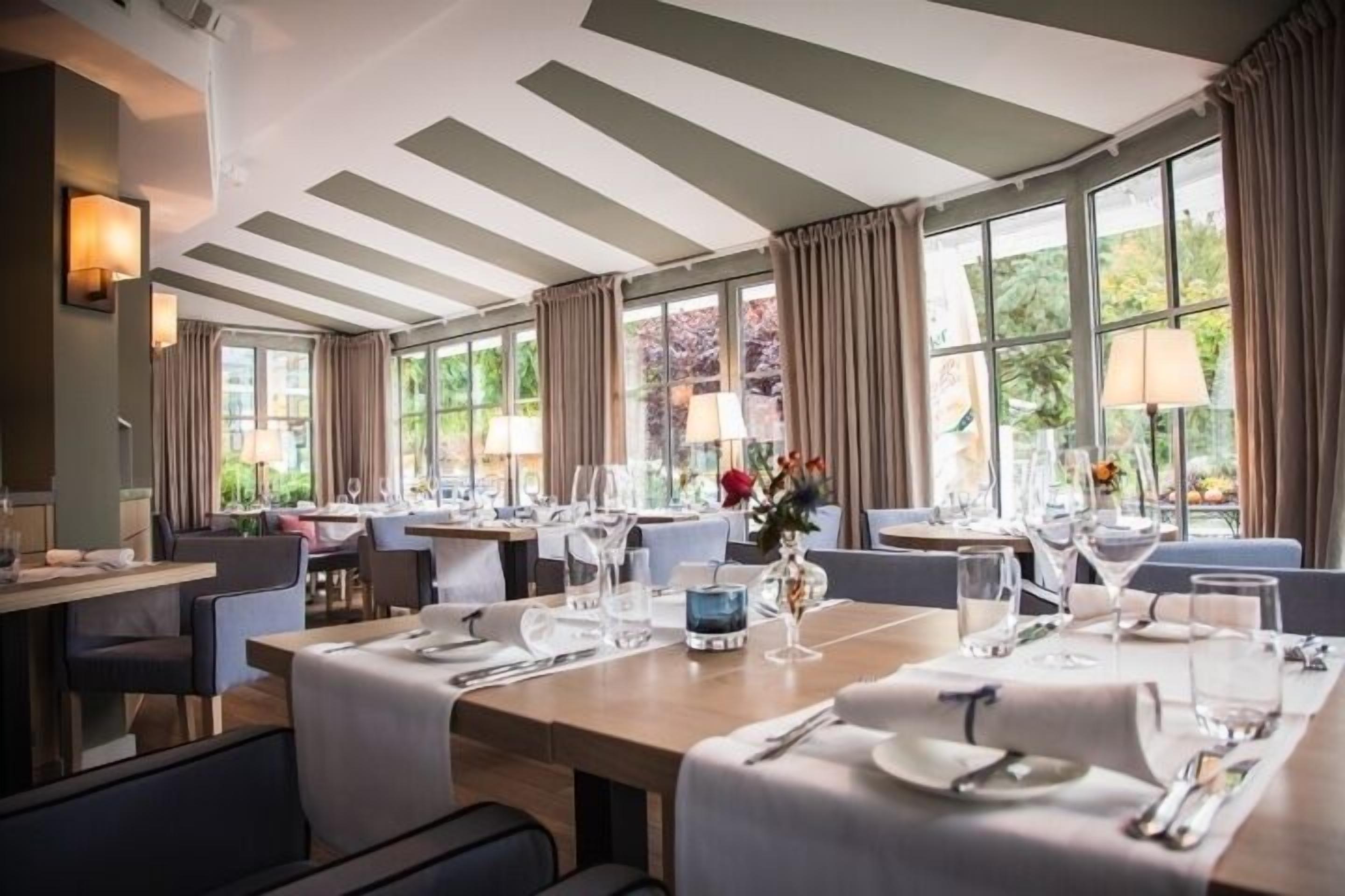 A dining area with tables set for service, large windows with curtains, and a view of greenery outside.