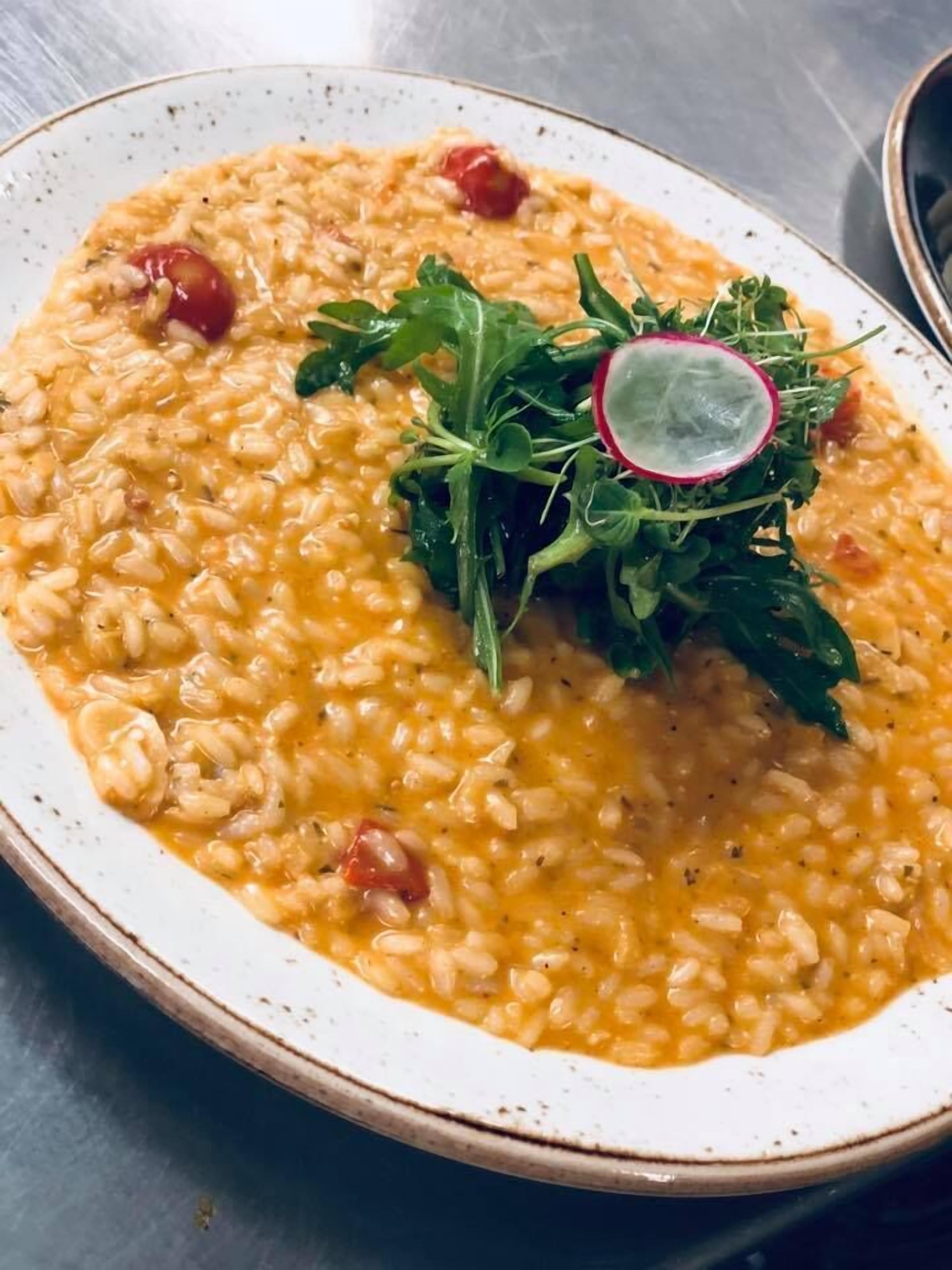 A bowl of risotto garnished with cherry tomatoes and a radish slice, served on a speckled plate.