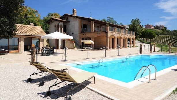Un espace piscine avec des chaises longues, un bâtiment doté d’un toit en tuiles et, au loin, un vignoble.