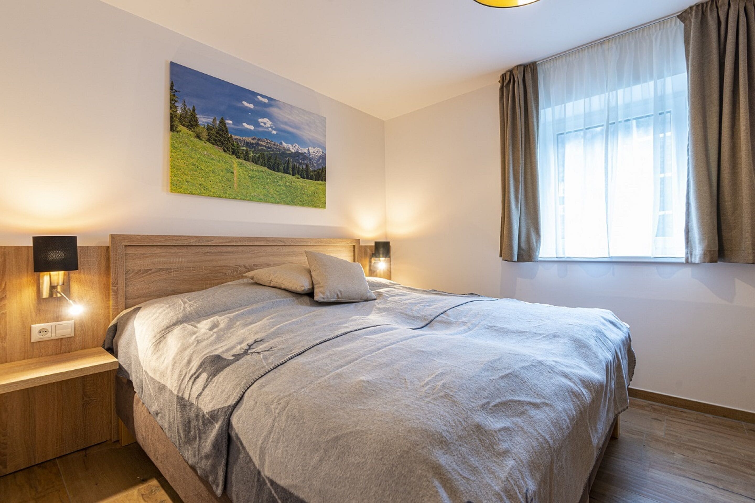 Quarto com cabeceira de madeira, cama grande com manta cinzenta, duas luminárias de cabeceira, mesa de cabeceira, janela com cortinas e um quadro de paisagem na parede.
