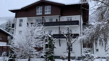 Un edificio de varios pisos con fachada de madera y paredes blancas, rodeado de árboles cubiertos de nieve y un telón de fondo montañoso.
