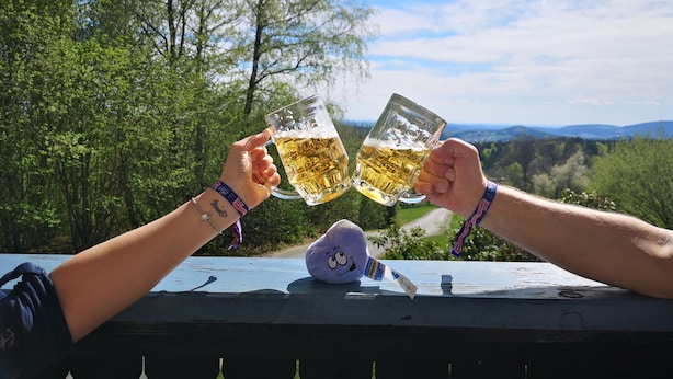 Two people toasting with beer mugs on a wooden deck with a scenic view of trees and hills in the background. - Pension Fernblick (Sankt Oswald-Riedlhuette)