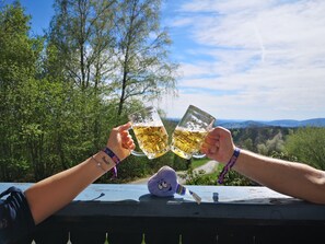 Two people toasting with beer mugs on a wooden deck with a scenic view of trees and hills in the background. - Pension Fernblick (Sankt Oswald-Riedlhuette)