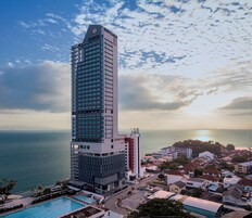 A tall hotel with a modern design, featuring a prominent clock on the top, a swimming pool, and a view of the sea.