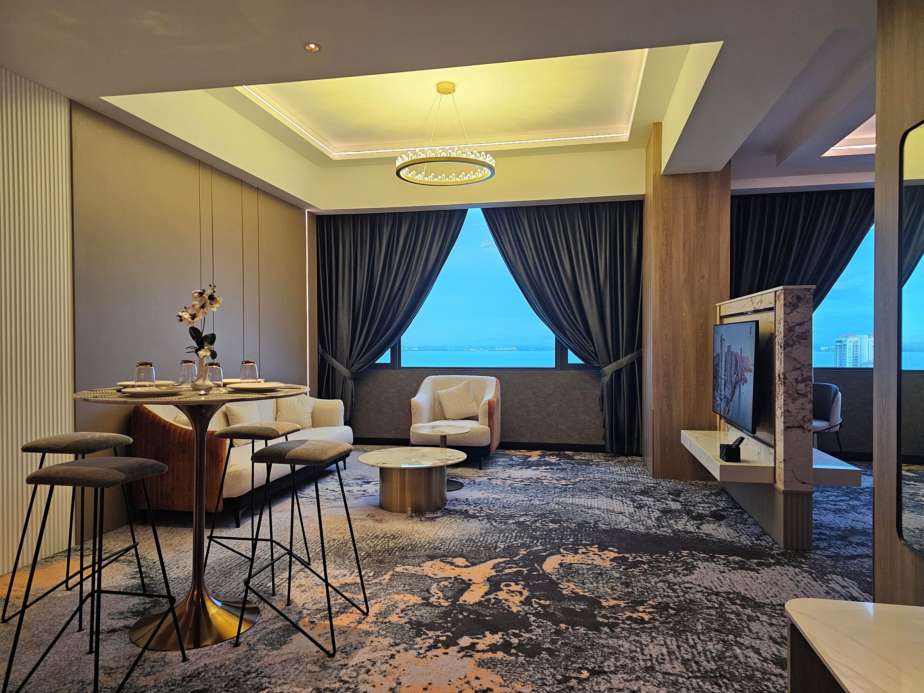 A modern hotel room with a dining area, a seating area with a view of the sea, and a television.