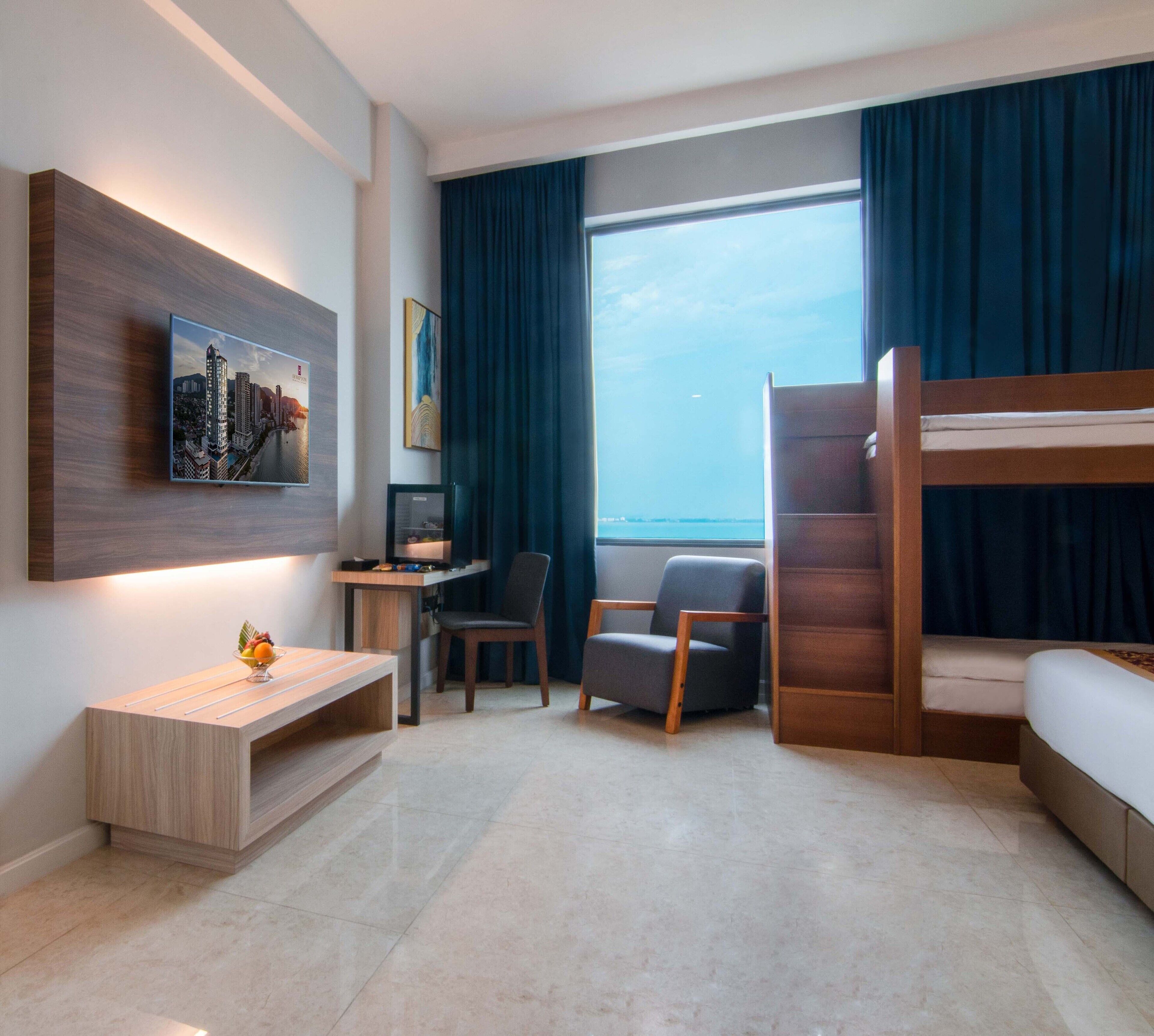 A modern hotel room with a bed, desk, chairs, and a large window with a view of the sea.