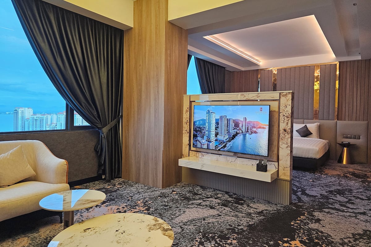 A modern hotel room with a large TV mounted on a wooden panel, a sofa, a coffee table, and a view of the cityscape.
