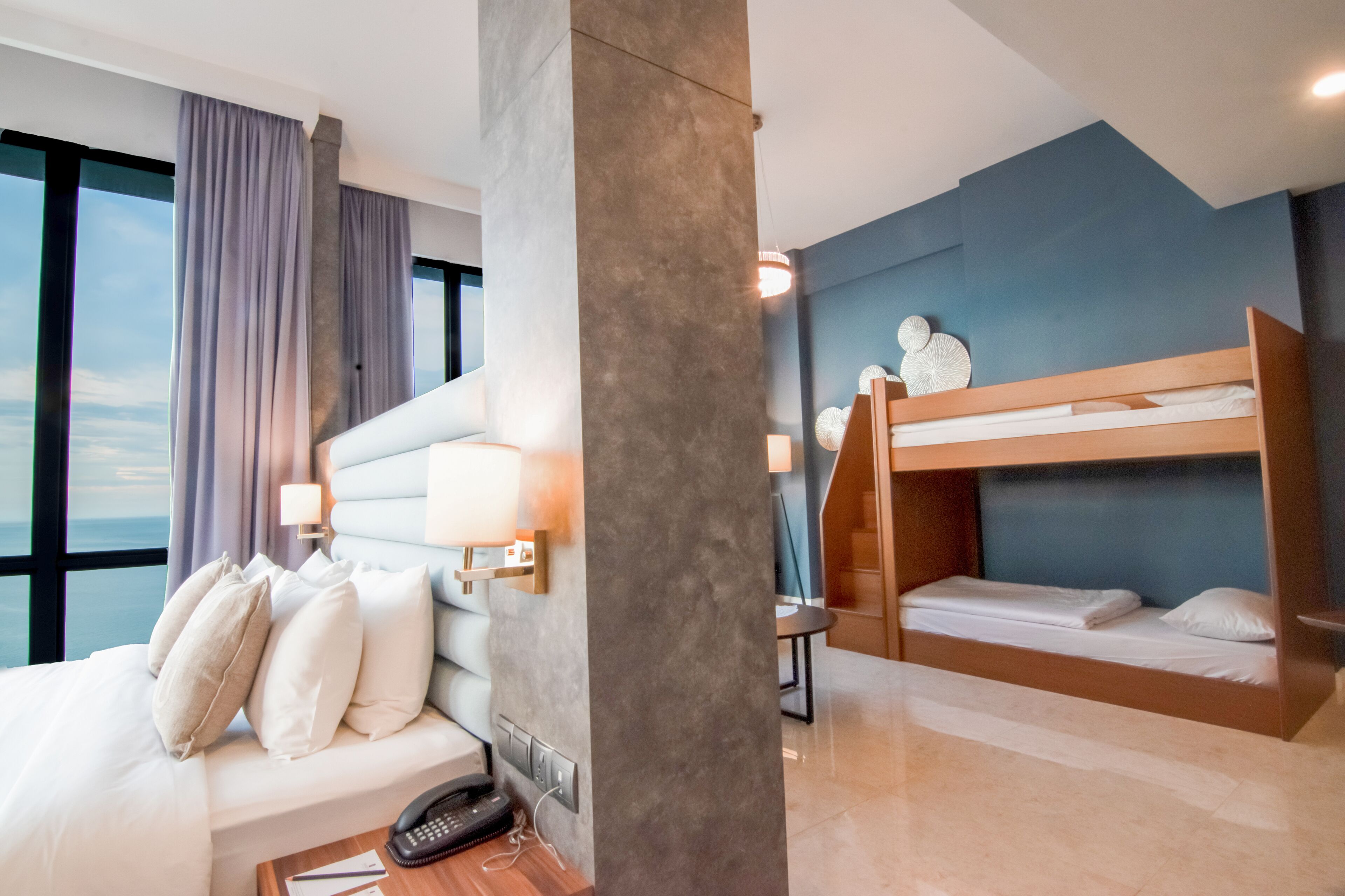 Habitación de hotel con una cama grande, una litera, un escritorio con teléfono y vistas al mar a través de amplias ventanas.