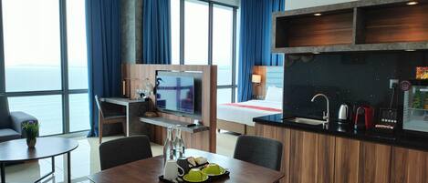 Habitación de hotel moderna con zona de comedor, kitchenette, televisor y un amplio ventanal con vistas al mar.