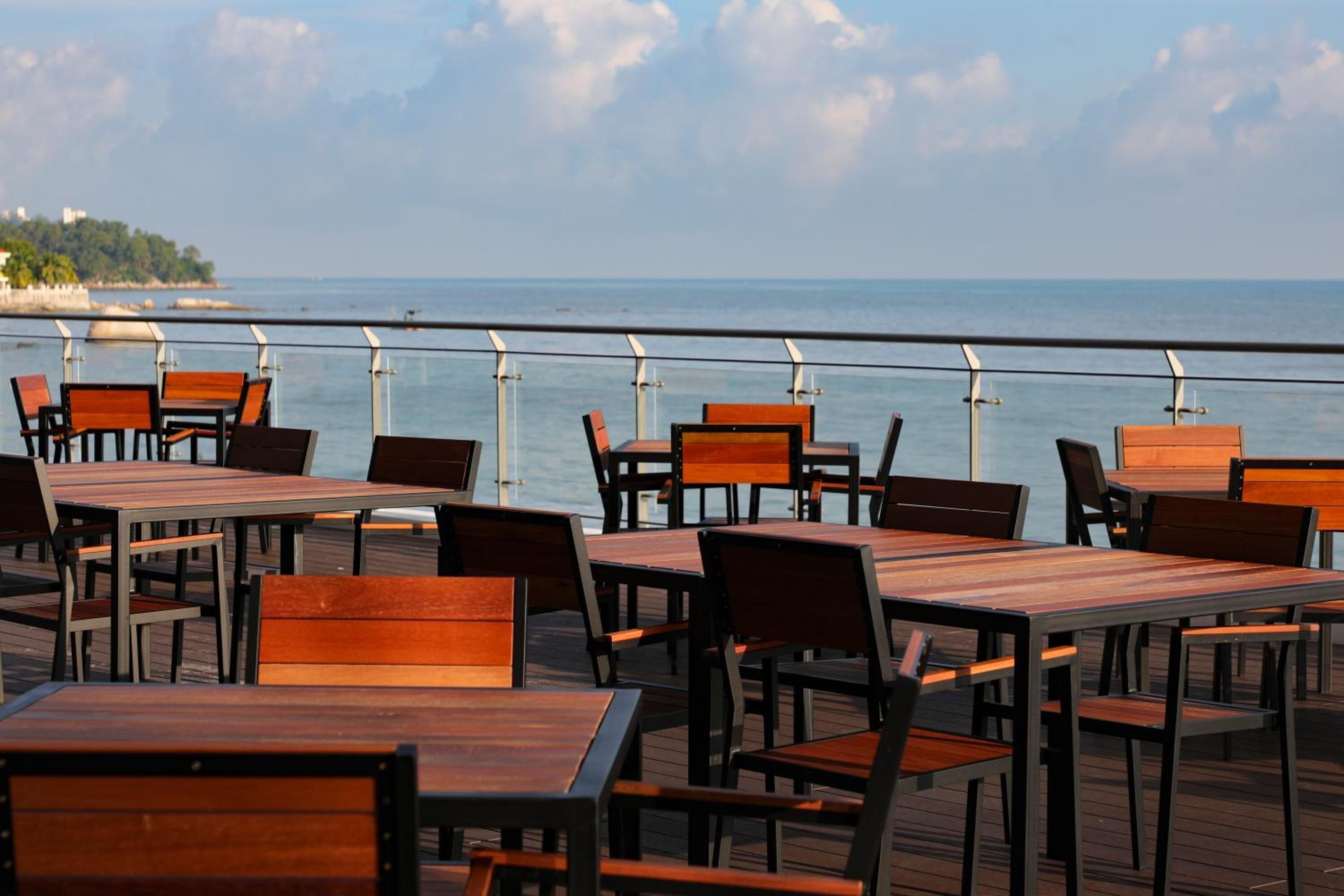 A deck with wooden tables and chairs, overlooking a body of water and a distant shoreline.