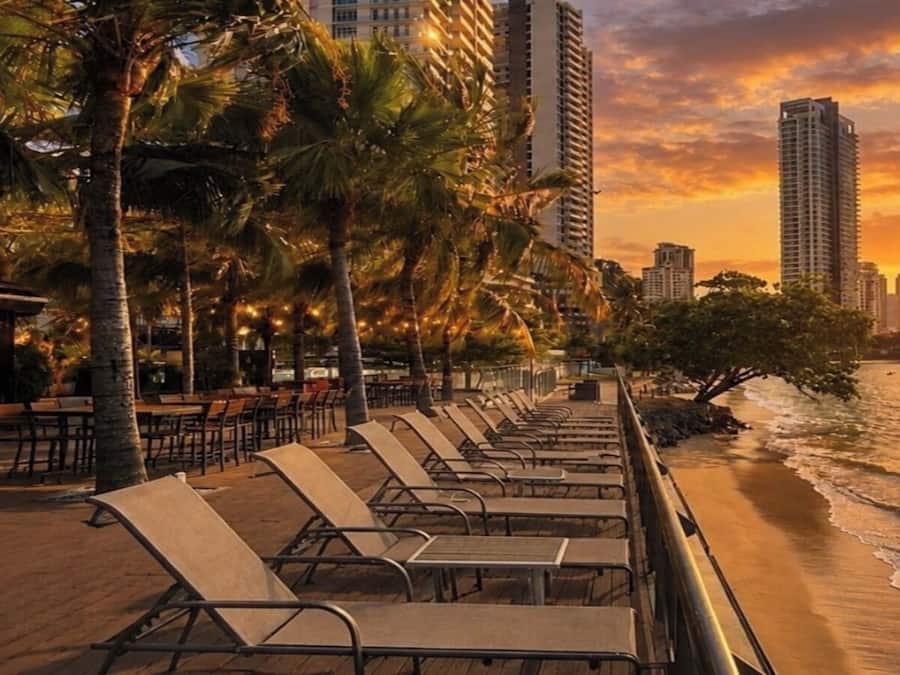 Un área de descanso frente a la playa con filas de sillones y mesas, palmeras y vistas al océano y al horizonte de la ciudad.