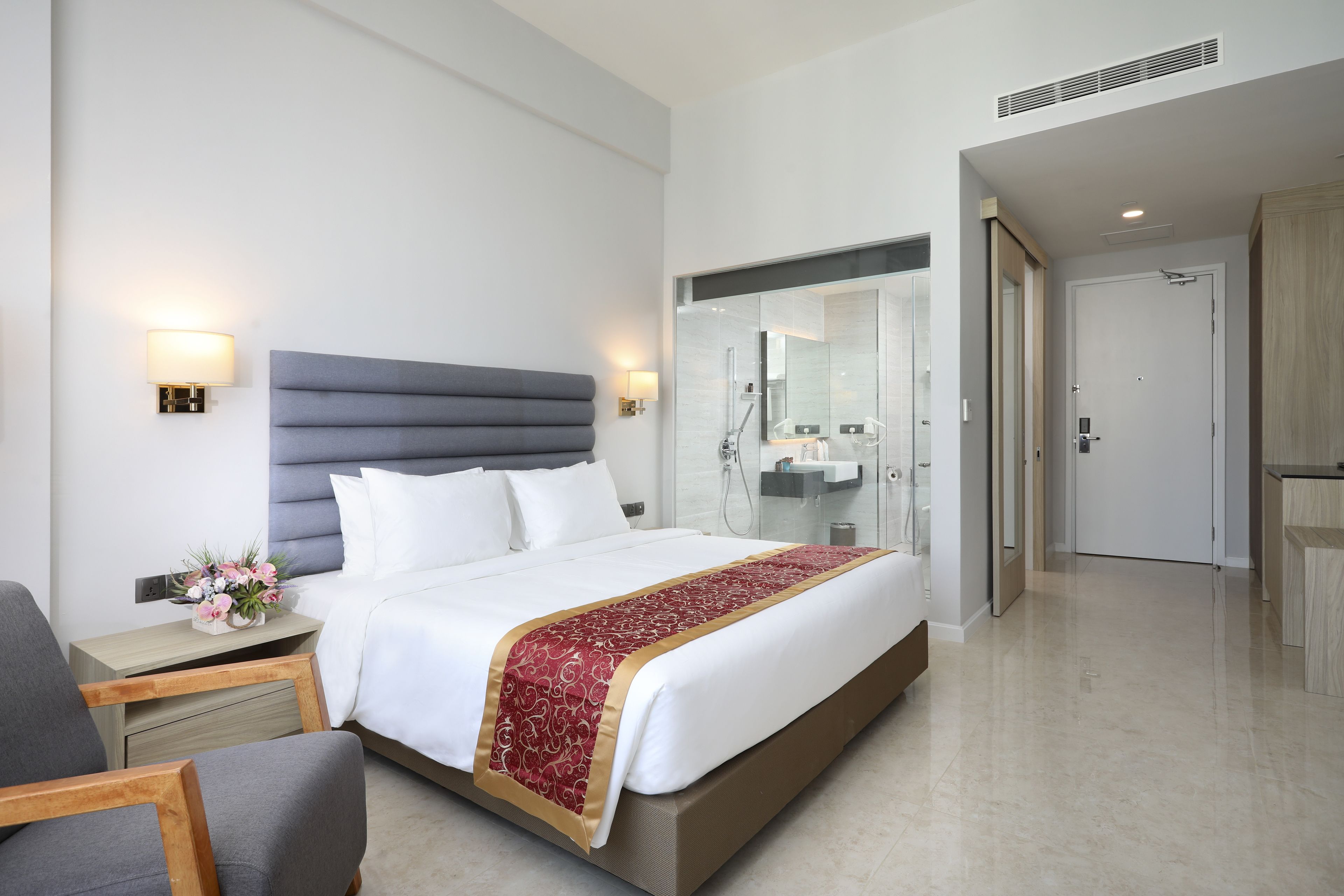 Une chambre d’hôtel moderne avec un grand lit, des tables de chevet, une petite table, un miroir et une salle de bain visible à travers une porte ouverte.