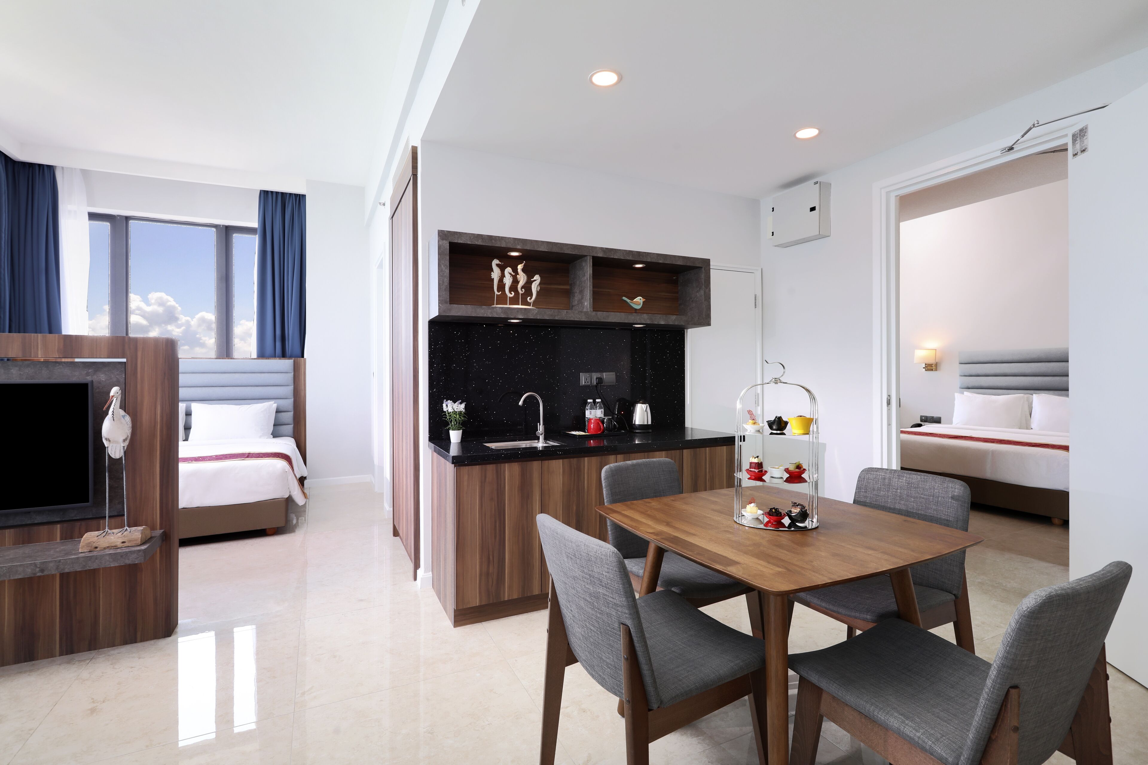 A modern hotel room with a wooden dining table, grey upholstered chairs, a kitchen area with a black countertop, and a large window with curtains.