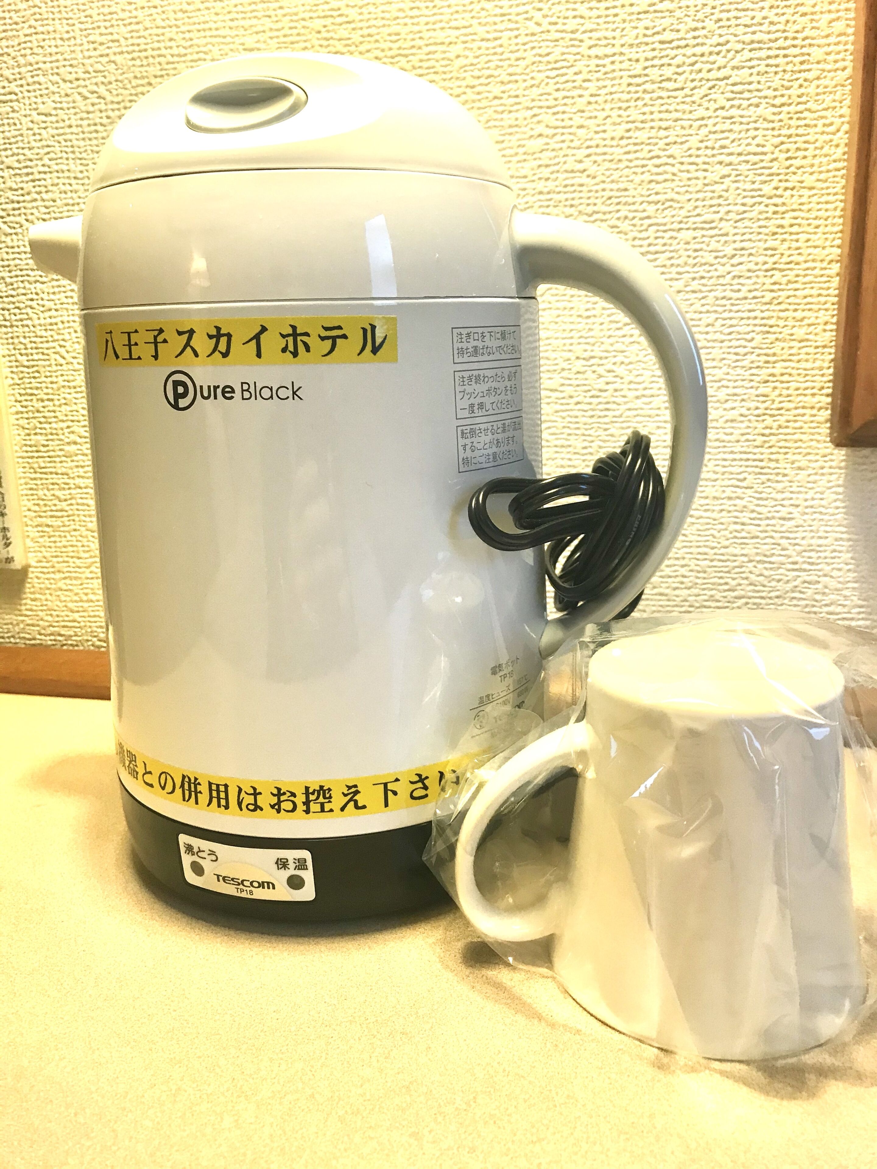 A white electric kettle with a handle and a cup attached to it, placed on a countertop.