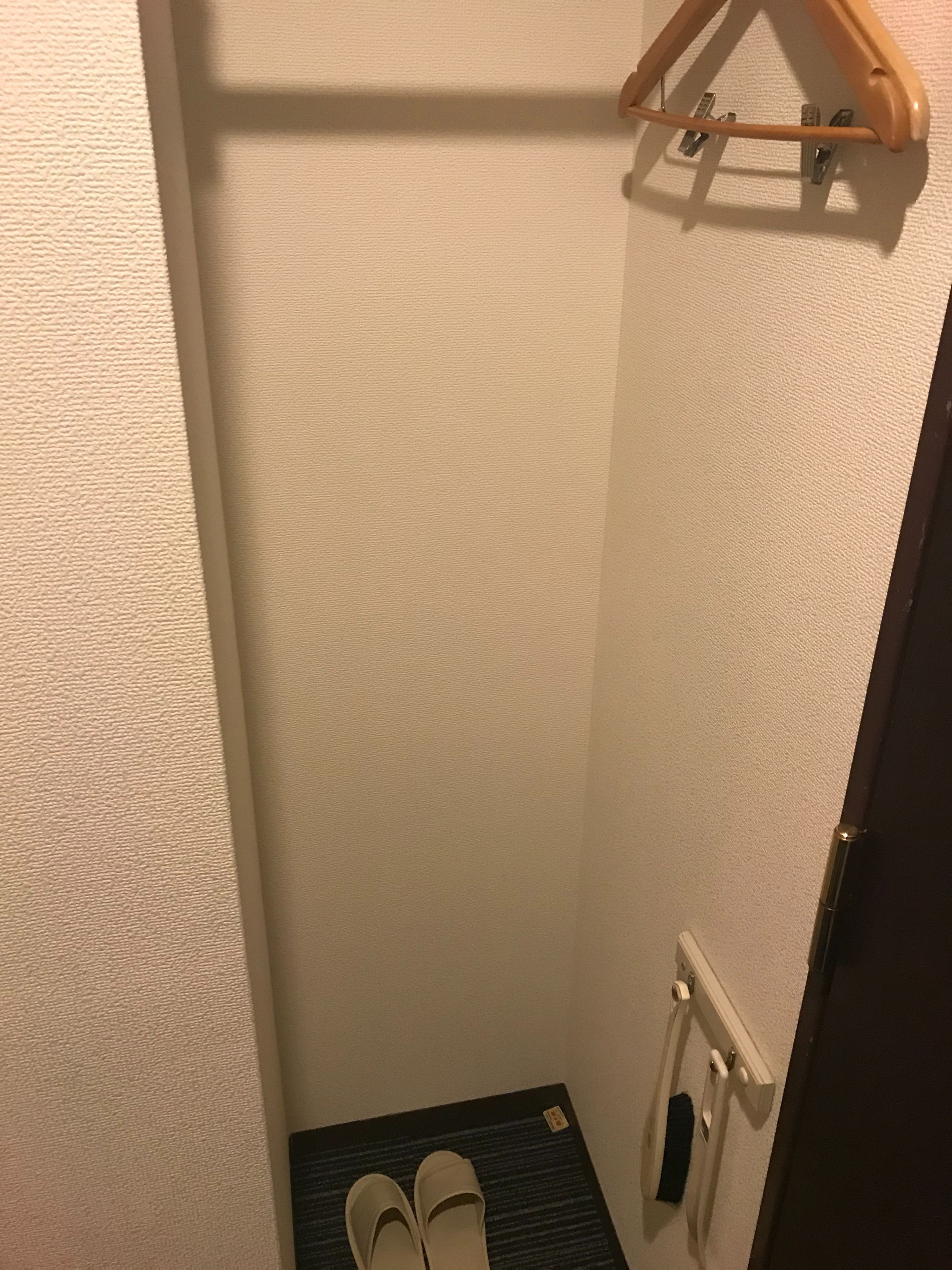 Una habitación pequeña con una pared blanca texturizada, un perchero de madera, un par de pantuflas blancas sobre un tapete negro y un radiador blanco.