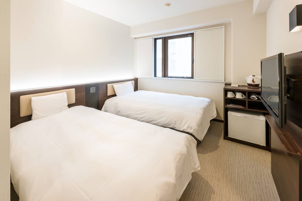 A hotel room with two beds, a television, a small refrigerator, and a window with blinds.