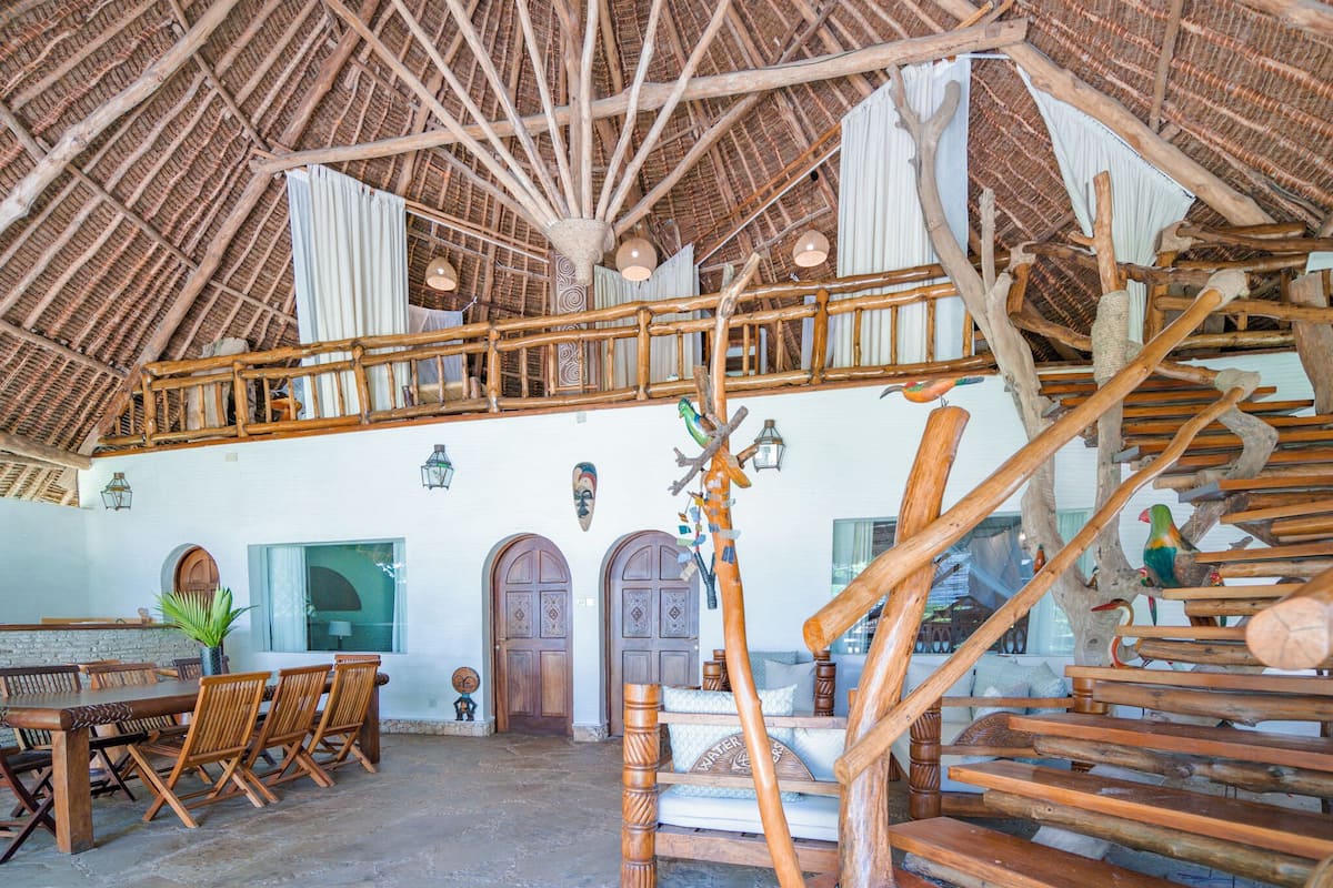 A rustic interior with a high wooden ceiling, a wooden staircase, and a dining area with wooden chairs and a table.