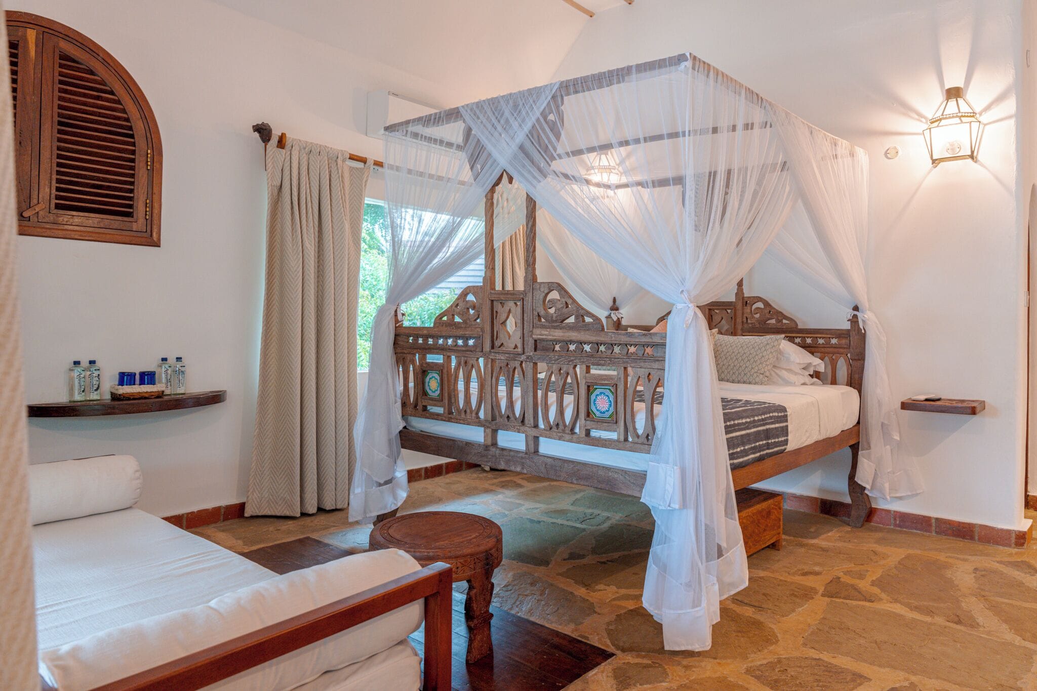 A room with a four-poster bed, a wooden bench, a small table, and a shelf with toiletries.