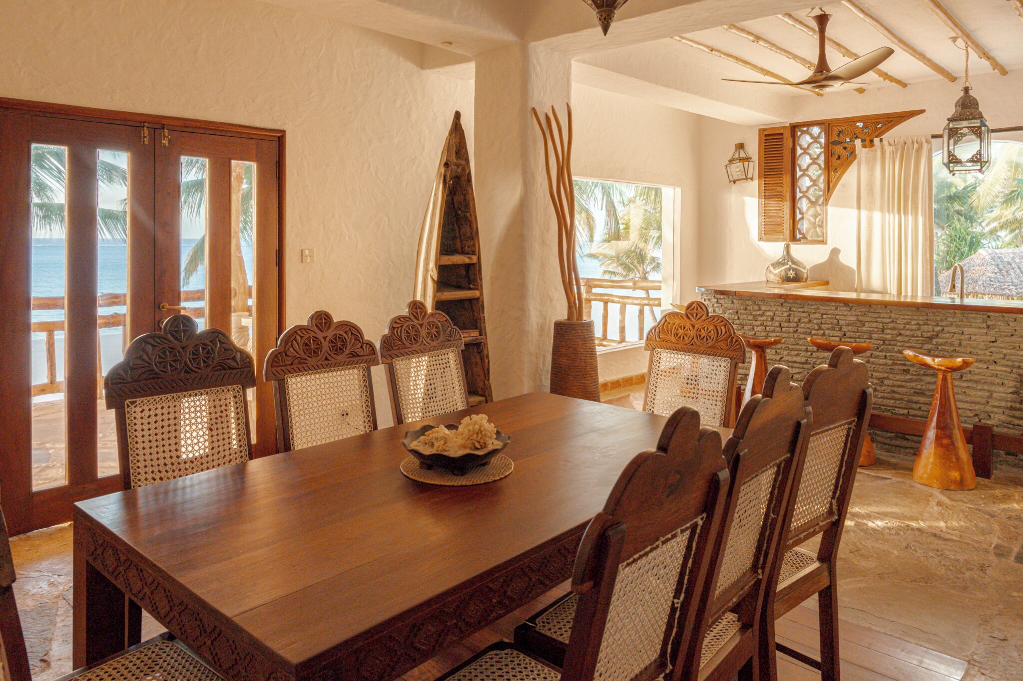 A wooden dining table with six chairs, a decorative centerpiece, and a view of the ocean through a wooden door.