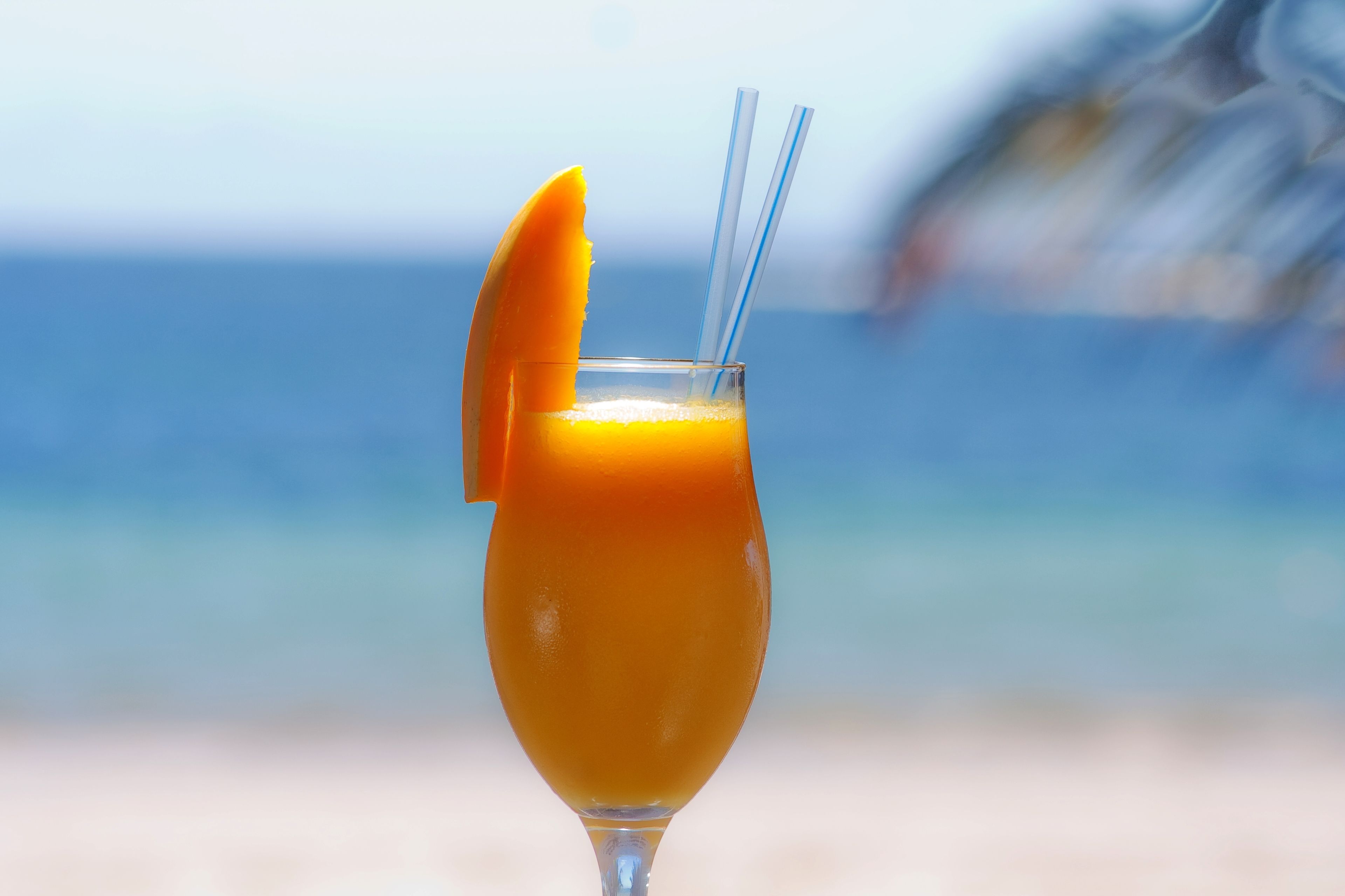 Un vaso de jugo de naranja con una rodaja de naranja y dos pajitas sobre una playa, con una vista borrosa del océano al fondo.
