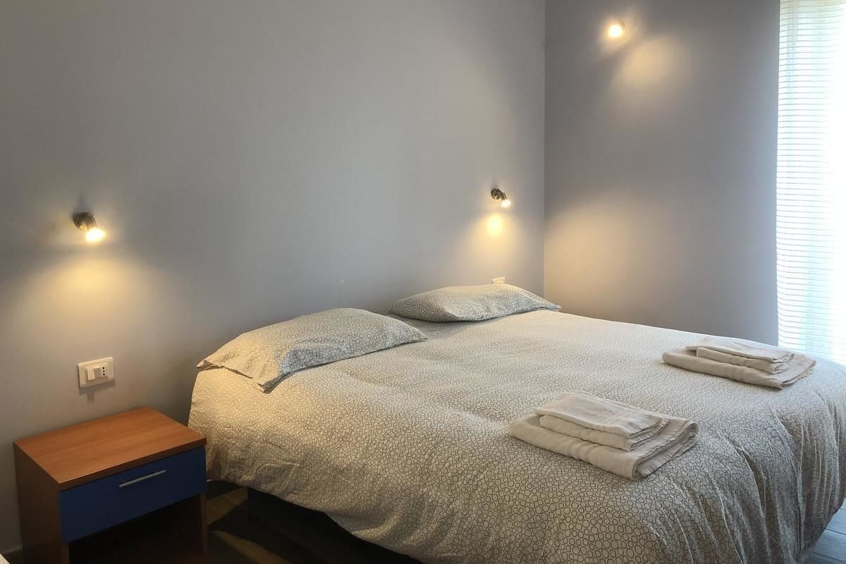 A neatly made bed with a patterned bedspread and pillows, a small wooden nightstand with a drawer, and two wall-mounted lights.