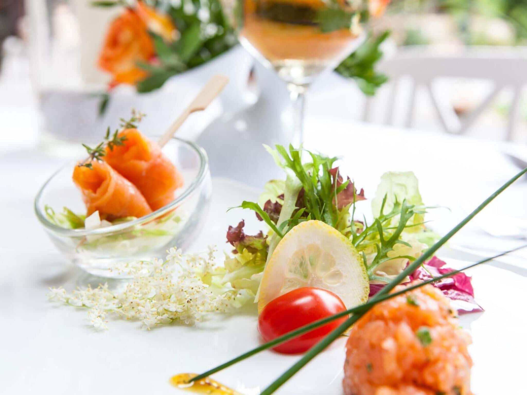 Une table dressée avec un verre contenant une boisson, un bol de saumon fumé, une salade garnie d’une tranche de citron et une petite assiette contenant ce qui ressemble à une tarte ou une pâte à tartiner.