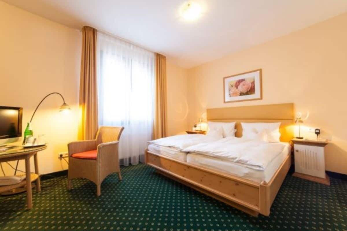 A hotel room with a bed, a desk with a chair, a radiator, and a window with curtains.