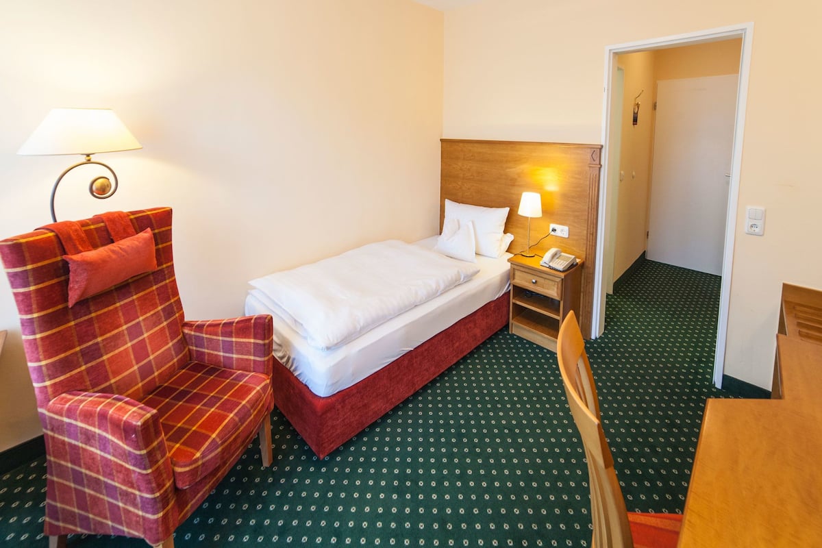 A hotel room with a bed, a chair, a nightstand with a lamp, and a telephone.