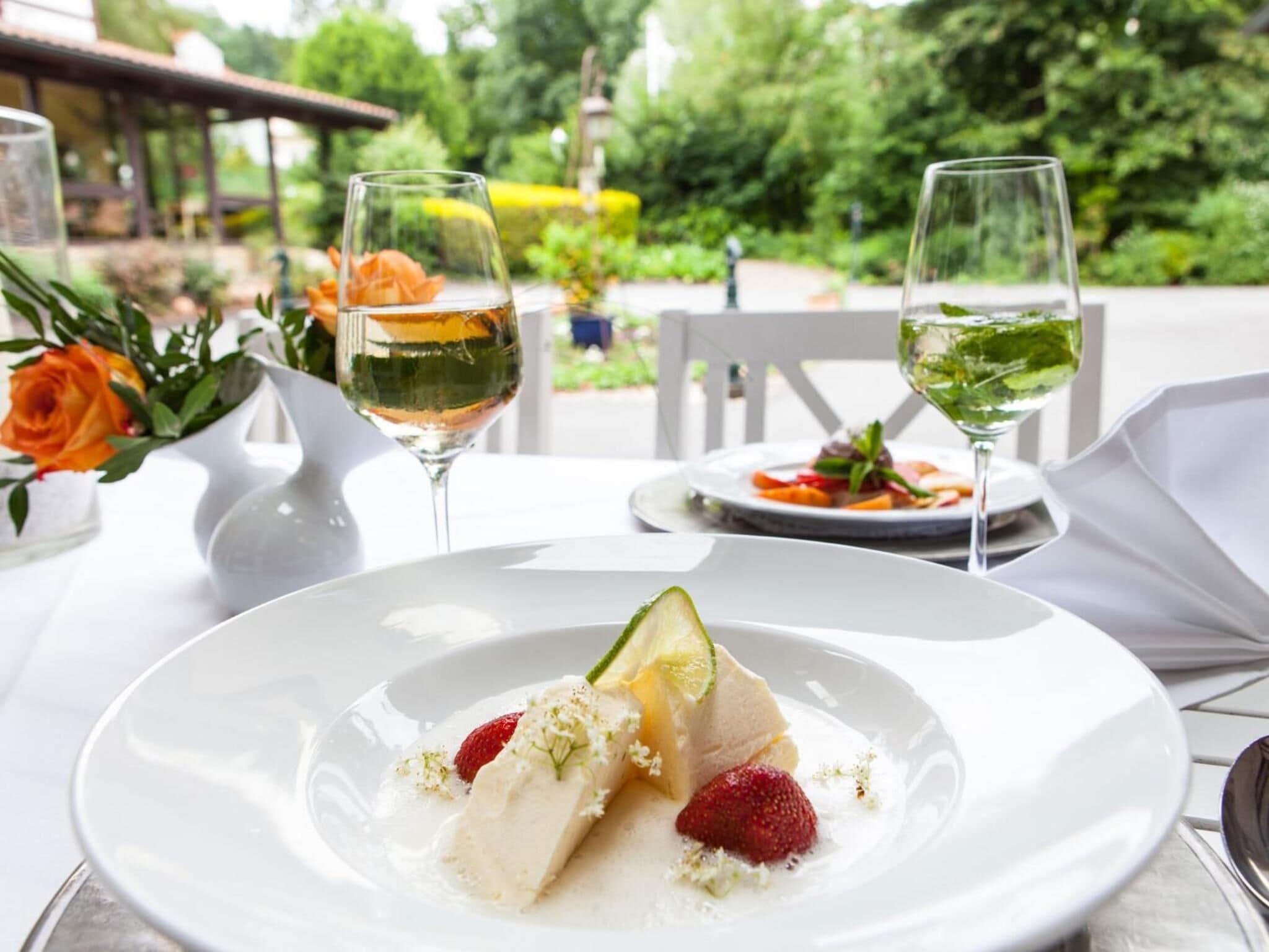 Una mesa dispuesta para una comida, con un plato de postre, dos copas de vino blanco y un jarrón con flores.