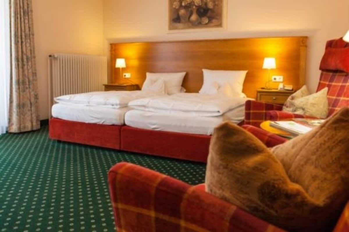 A hotel room with a double bed, two bedside lamps, a red plaid armchair, a small table with a book, and a painting on the wall.