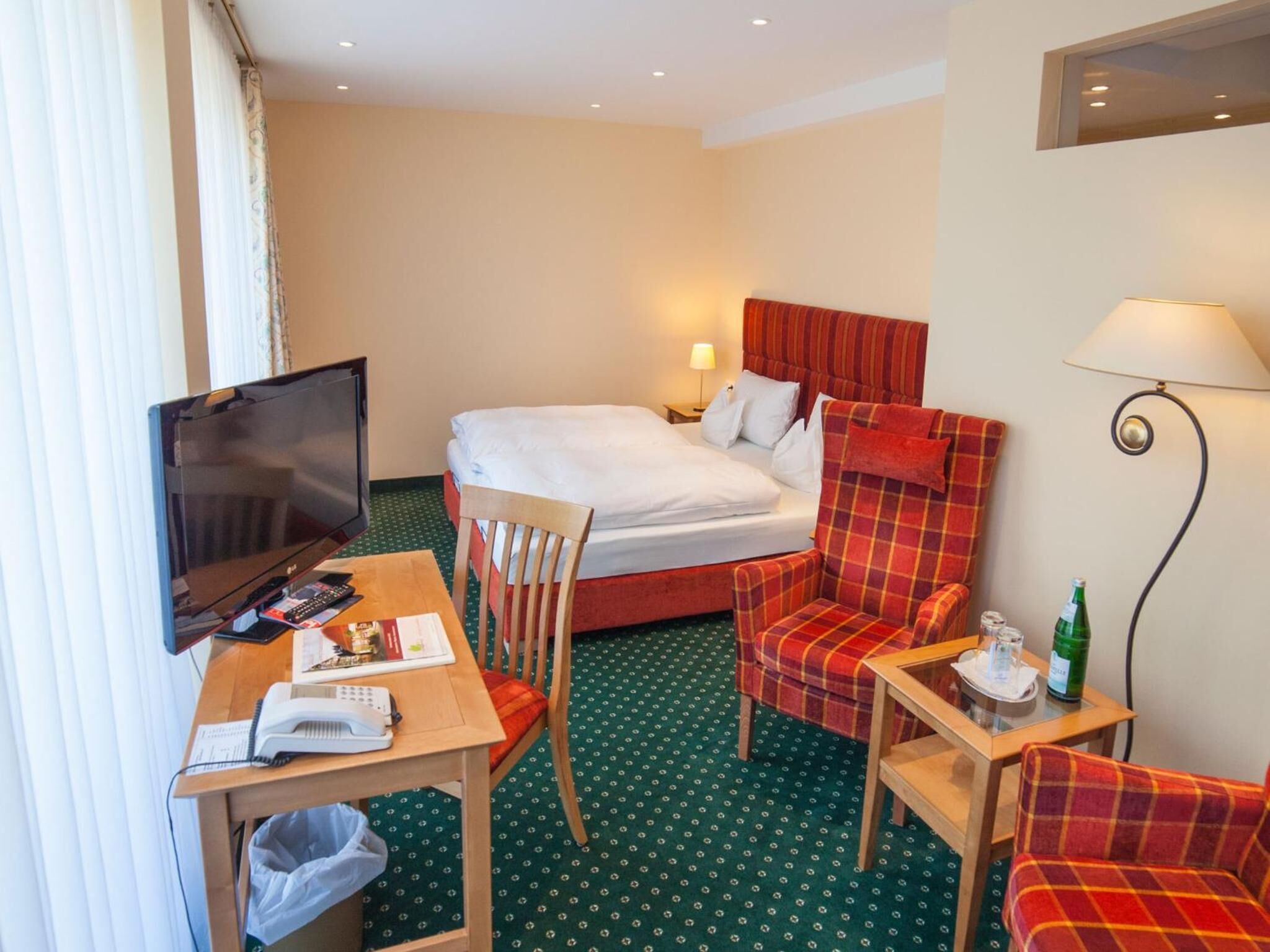 Ein Hotelzimmer mit einem Bett, einem Schreibtisch mit Fernseher, einem Sessel, einem kleinen Tisch mit Lampe und einer Couch.