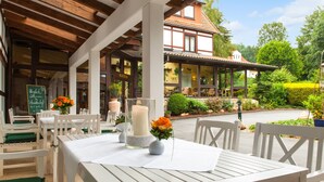 Un espace de restauration en plein air avec des tables et des chaises en bois peintes en blanc, un centre de table composé de bougies et une vue sur un bâtiment traditionnel doté d’un toit en tuiles.