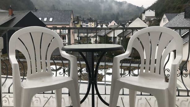 Two white plastic chairs and a small round table on a balcony with a view of a residential area and hills in the background. - Ferienwohnungen Domizil (Bad Bertrich)